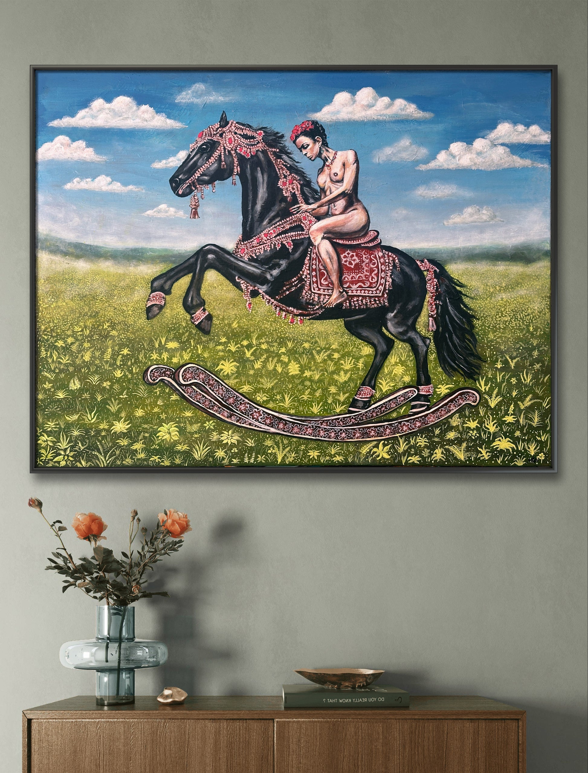 Framed artwork of a woman riding a horse in a field with a vase and books on a wooden cabinet below.